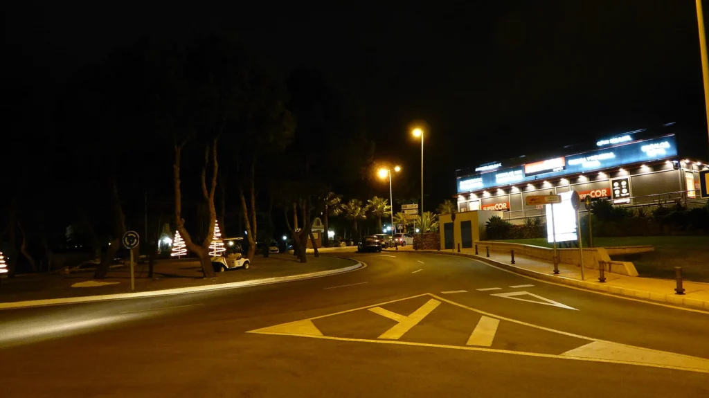 Rotonda y calle de noche iluminadas por farolas, con un edificio de luces azules al fondo y el asfalto con reflejos