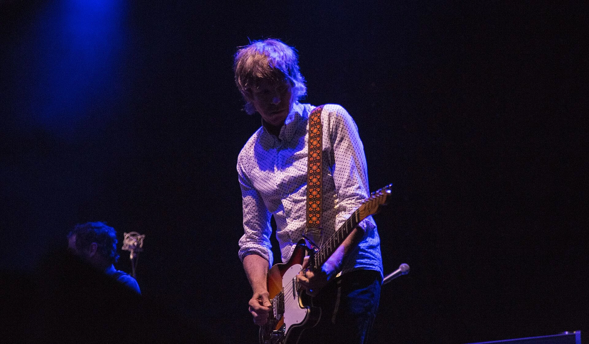 Patrick Sansone tocando la guitarra eléctrica en un escenario con luz azul de fondo