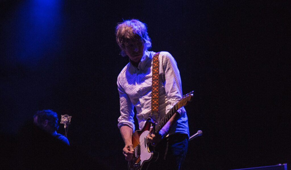 Patrick Sansone tocando la guitarra en un concierto, iluminado sobre fondo oscuro
