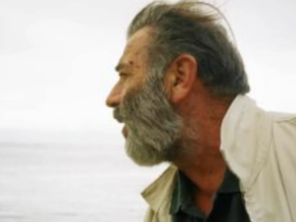 Vari Caramés de perfil, con barba canosa y chaqueta clara, frente al mar en un día gris
